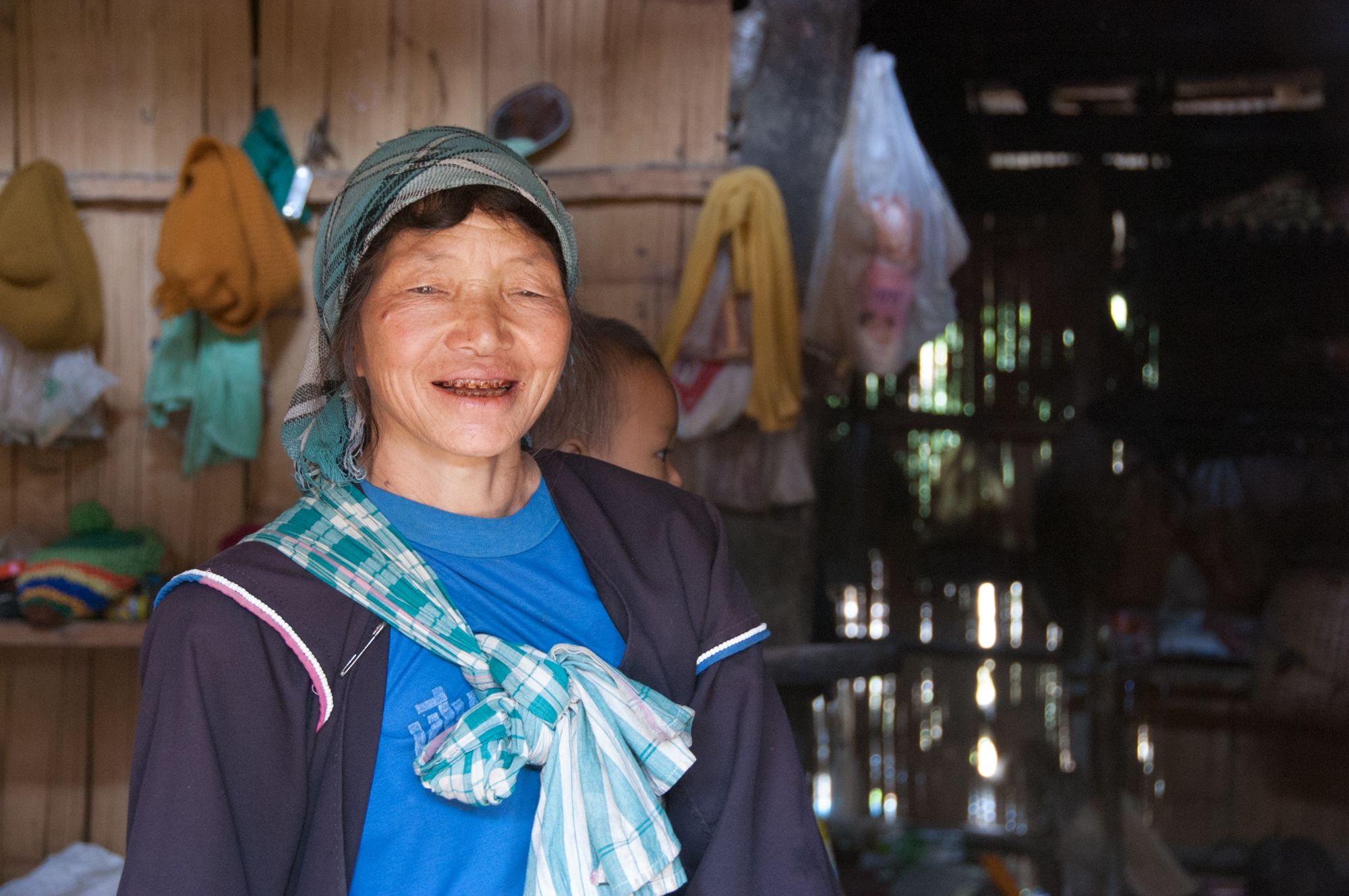 A woman carries and infant on her back. She stands inside a small wooden hut that is the family home.